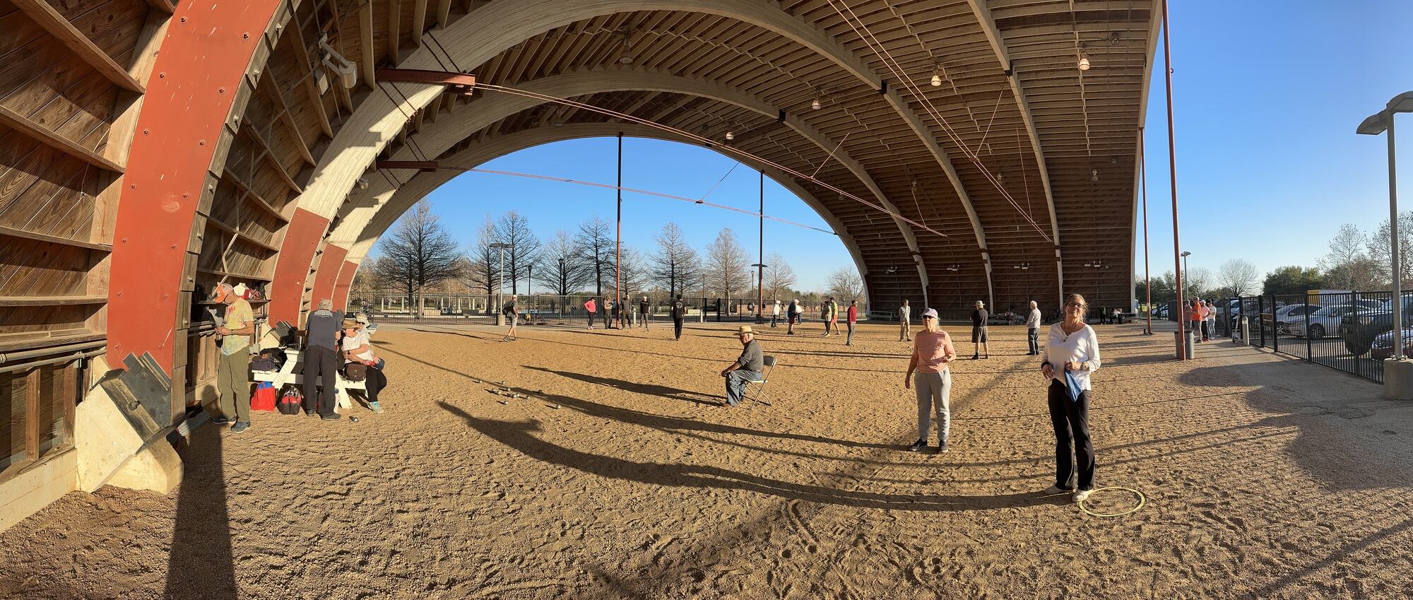 Panorama under the arches on a sunny afternoon: people spread across the open gravel floor, some playing, one sitting in a folding chair, a few at a picnic table by the wooden wall.