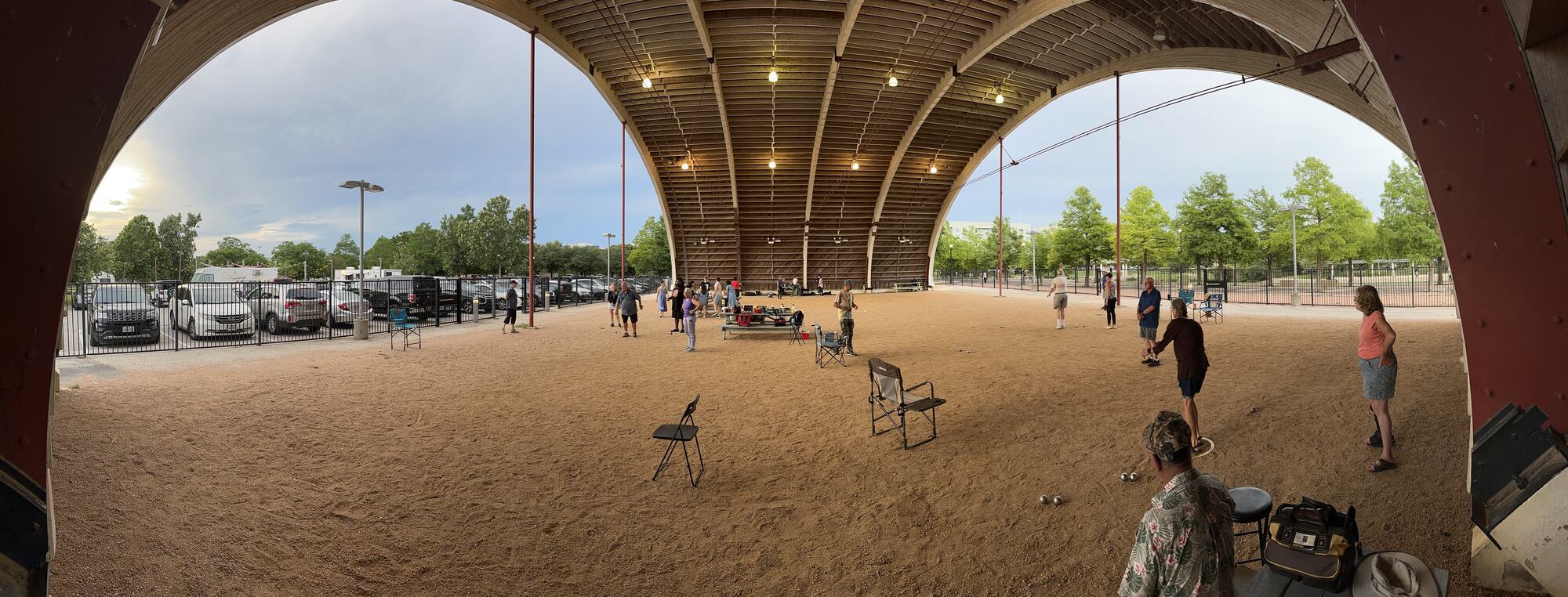 Wide view under the Hangar's wooden arches on a weekend afternoon: people playing pétanque on the open gravel floor, folding chairs, trees and sky at both open ends.